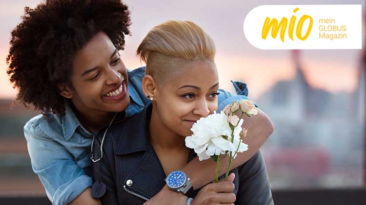 Person in schwarzer Jacke hält einen Strauß weißer und rosafarbener Blumen, Schriftzug „mio mein GLOBUS Magazin“ oben rechts.