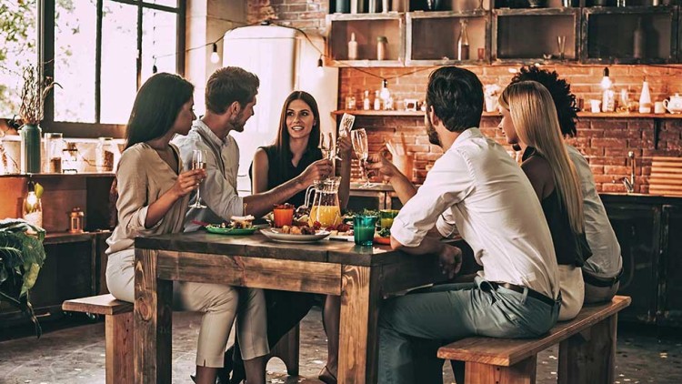 Sechs Personen sitzen an einem Holztisch in einer Küche und stoßen mit Gläsern an, auf dem Tisch stehen Speisen und Getränke.