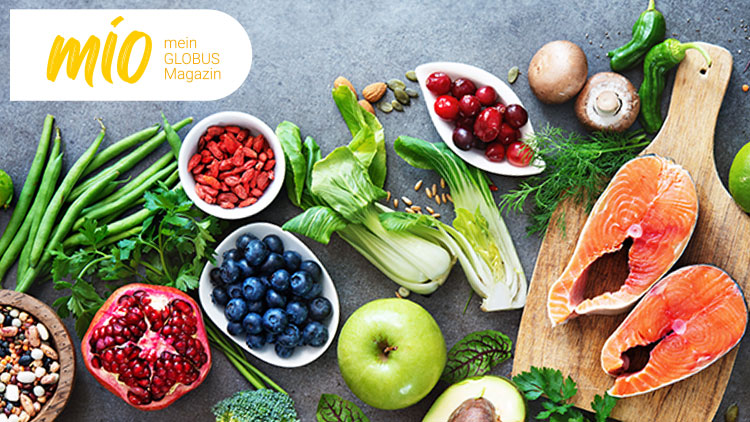 Bunte Auswahl an frischen Lebensmitteln wie Obst, Gemüse, Samen, Hülsenfrüchten und Lachsfilets auf grauer Oberfläche, oben mio mein GLOBUS Magazin.