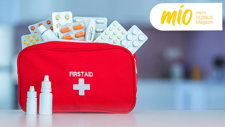 Rote Erste-Hilfe-Tasche mit der Aufschrift „FIRST AID“ und weißem Kreuz, gefüllt mit Medikamenten und zwei kleinen Medizinflaschen.