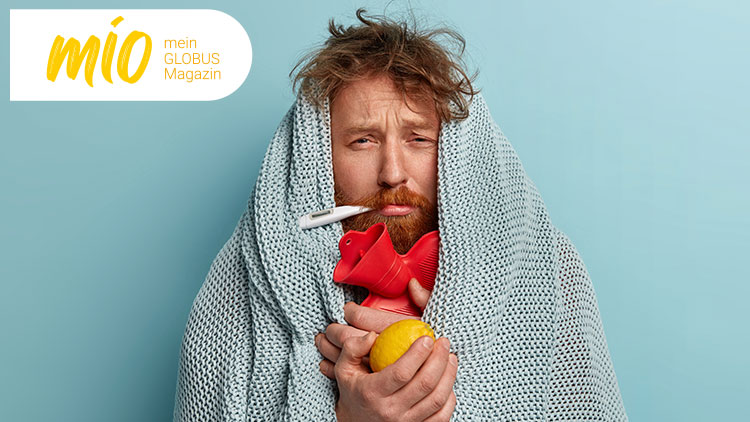 Person in eine graue Strickdecke gehüllt hält eine rote Wärmflasche und eine gelbe Zitrone vor einem hellblauen Hintergrund, mio mein GLOBUS Magazin-Logo oben links.