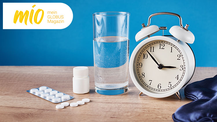 Ein weißer Wecker, ein Glas Wasser, Tabletten, eine Dose und eine blaue Schlafmaske auf einem Holztisch vor blauem Hintergrund.