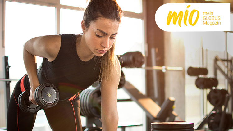 Person im schwarzen Sportoutfit trainiert mit einer Hantel in einem Fitnessstudio, rechts oben steht „mio mein GLOBUS Magazin“.