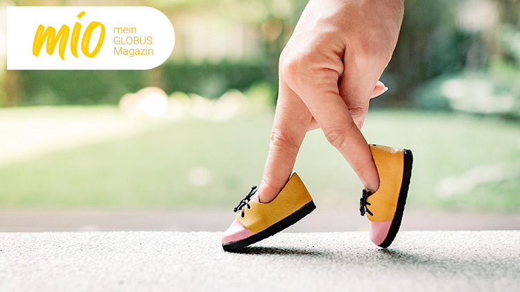 Eine Hand mit Fingern in gelb-pinken Schuhen läuft auf einer Steinfläche, Text oben links: mio mein GLOBUS Magazin.