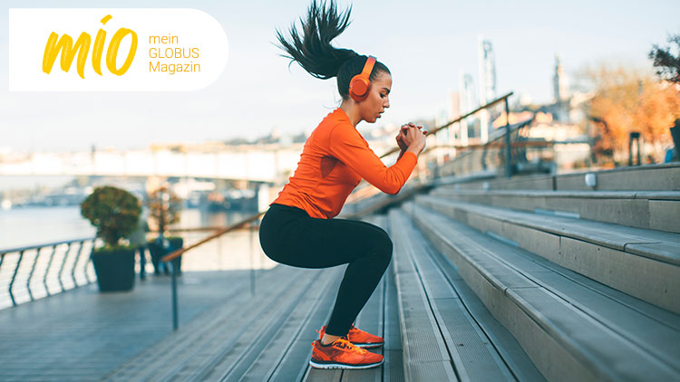 Person in orange Pullover macht Sport auf Treppen mit Kopfhörern, im Hintergrund ein Fluss, Bäume und eine Brücke.