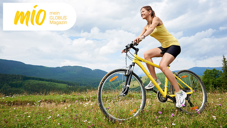 Person fährt auf gelbem Fahrrad durch eine Wiesenlandschaft vor bewaldeten Hügeln, oben links steht „mio, mein GLOBUS Magazin“.