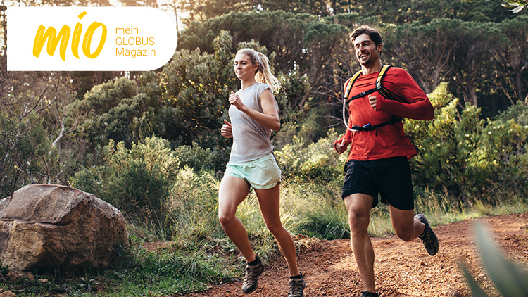 Zwei Personen joggen auf einem Waldweg, links im Bild ist ein großer Stein, oben links steht „mio mein GLOBUS Magazin“.