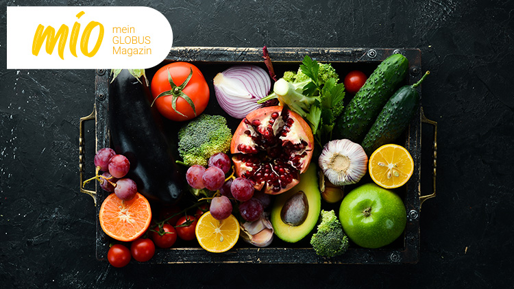 Bunte Auswahl an frischem Obst und Gemüse, darunter Granatapfel, Brokkoli, Gurken, Tomaten, Avocado, Trauben und Zitrusfrüchte, daneben mio mein GLOBUS Magazin.