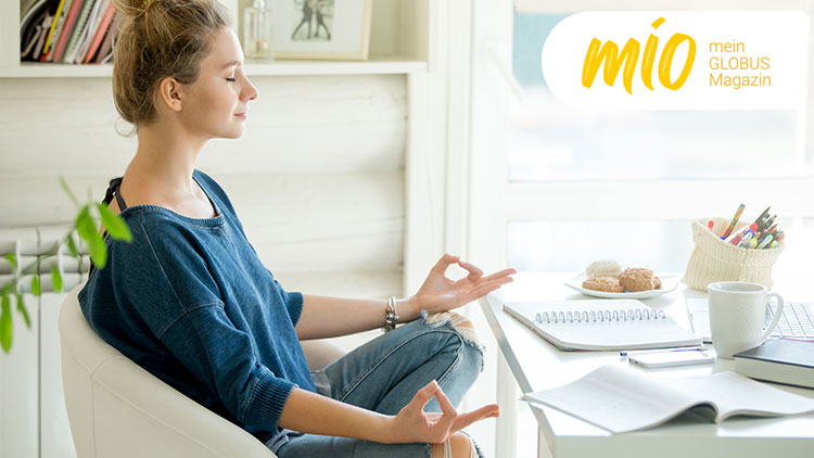 Person sitzt in Meditationshaltung an einem Tisch mit einer Karteikiste, Keksen, einer Tasse und dem Schriftzug mio mein GLOBUS Magazin.