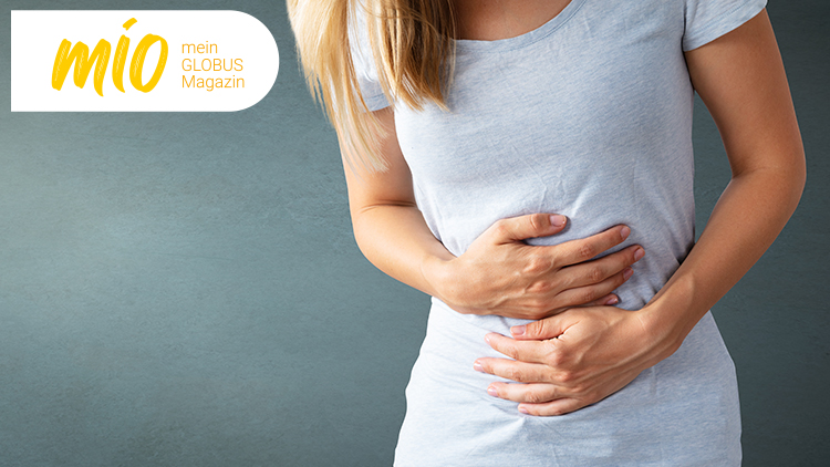 Person in grauem T-Shirt hält beide Hände auf den Bauch, links oben steht „mio mein GLOBUS Magazin“ in Gelb und Orange.