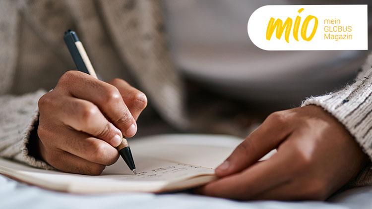 Eine Hand schreibt mit einem Stift in ein aufgeschlagenes Notizbuch, oben rechts steht „mio mein GLOBUS Magazin“ in Gelb und Weiß.