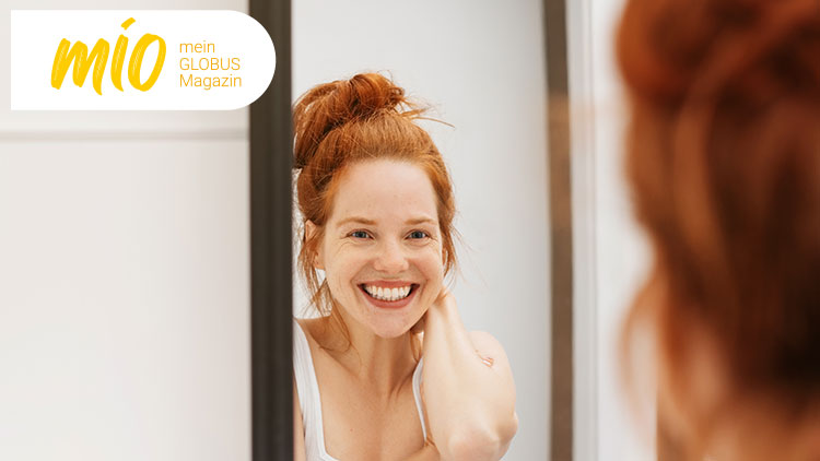 Eine Person mit hochgesteckten roten Haaren betrachtet sich in einem Spiegel, links oben steht „mio mein GLOBUS Magazin“.