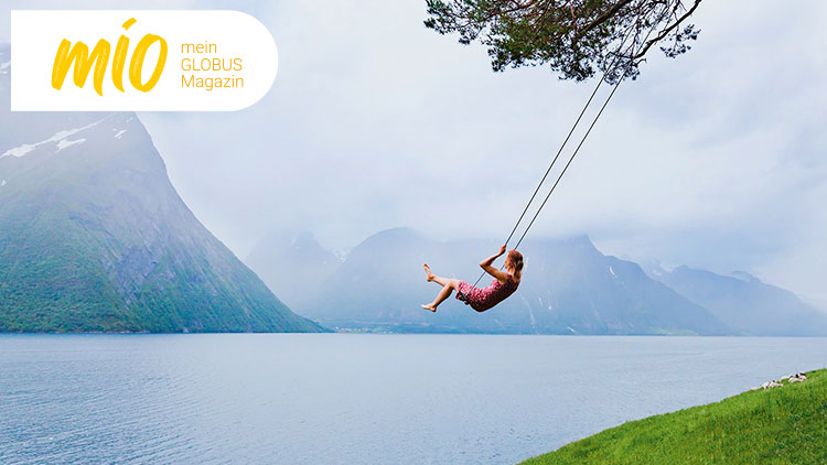 Eine Person schaukelt über einem See mit bergiger Landschaft, links oben das mio mein GLOBUS Magazin Logo in Gelb und Orange.
