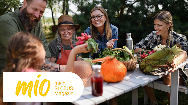 Eine Gruppe von Menschen sitzt an einem Tisch im Freien mit frischem Gemüse, im Vordergrund das mio mein GLOBUS Magazin-Logo.