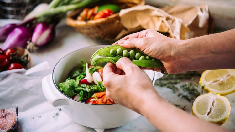 Hände öffnen eine grüne Erbsenschote über einer weißen Schüssel mit frischem Salat, im Hintergrund liegt Gemüse.