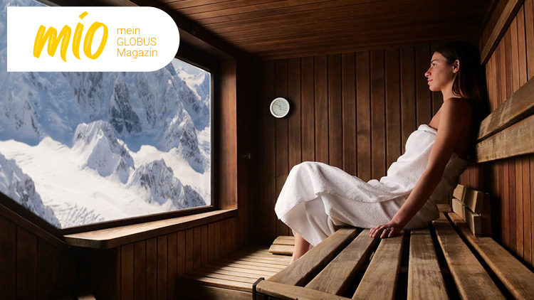 Person in einer hölzernen Sauna sitzt mit weißem Handtuch, durch ein Fenster ist eine winterliche Berglandschaft sichtbar, mio mein GLOBUS Magazin.