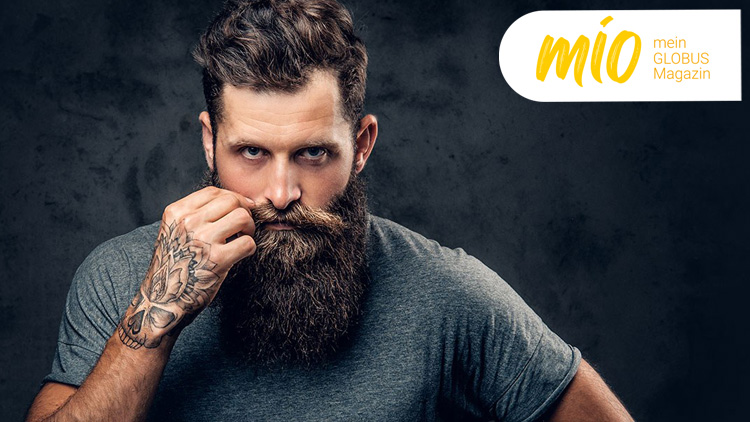 Person mit tätowierter Hand hält einen langen, dunklen Bart vor dunklem Hintergrund, „mio mein GLOBUS Magazin“-Logo oben rechts.