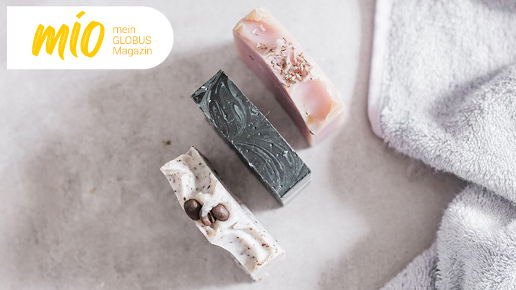 Drei Stücke Seife in Rosa, Schwarz und Weiß mit Verzierung liegen auf grauem Untergrund, daneben ein hellgraues Handtuch, oben links „mio mein GLOBUS Magazin“.