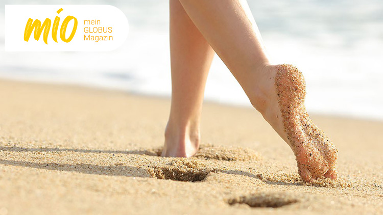 Ein nackter Fuß mit Sand geht über einen Strand, links oben steht „mio, mein GLOBUS Magazin“ in gelber und weißer Schrift.