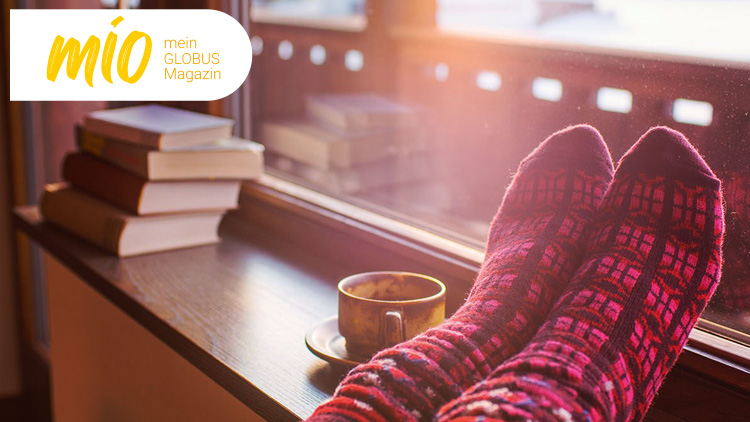 Eine Tasse Kaffee, rote Stricksocken und Bücher liegen auf einer Fensterbank mit Sonnenlicht, Text: mio, mein GLOBUS Magazin.
