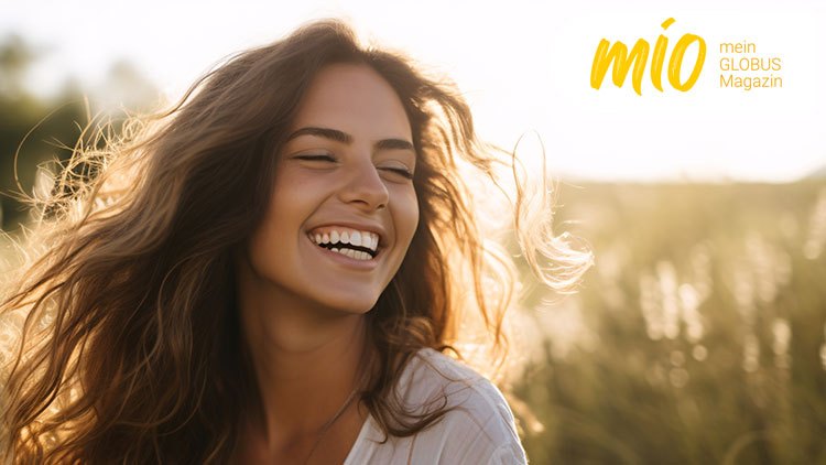 Eine Person mit langen, welligen Haaren steht auf einer Wiese bei Sonnenschein, rechts oben steht „mio mein GLOBUS Magazin“.