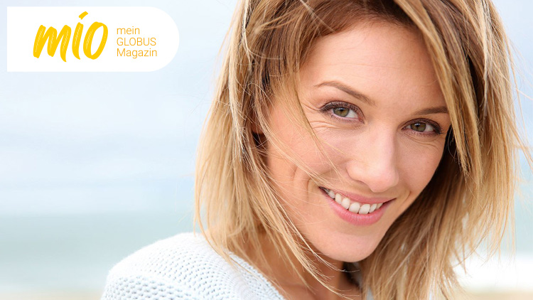 Blondes Haar im Wind, im Hintergrund ein heller Himmel und ein unscharfer Küstenhintergrund, oben links mio mein GLOBUS Magazin.