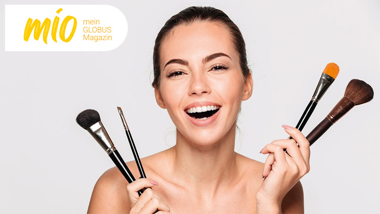 Eine Person hält vier Make-up-Pinsel vor einem hellen Hintergrund, links oben steht mio mein GLOBUS Magazin.