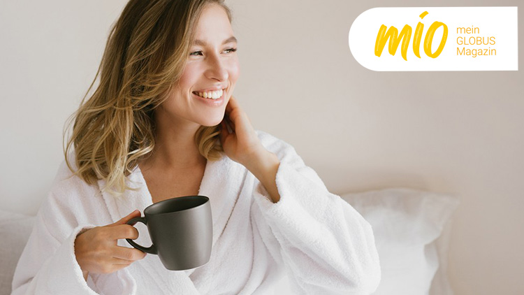 Person in weißem Bademantel sitzt mit grauer Tasse in der Hand auf einem Bett, oben links steht „mio mein GLOBUS Magazin“.