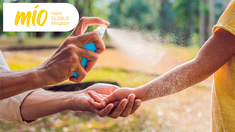 Eine Hand sprüht aus einer blauen Flasche Flüssigkeit auf einen Arm, oben links steht „mio mein GLOBUS Magazin“.