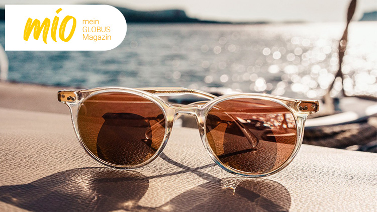 Eine Sonnenbrille mit transparentem Gestell und braunen Gläsern liegt auf einer hellbraunen Oberfläche vor verschwommenem Wasserhintergrund.