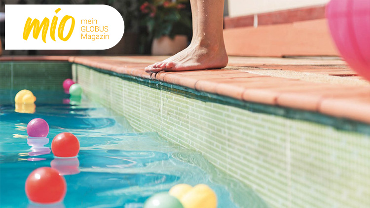 Ein nackter Fuß steht auf dem Rand eines Pools mit bunten Plastikbällen im Wasser, rechts oben steht „mio mein GLOBUS Magazin“.