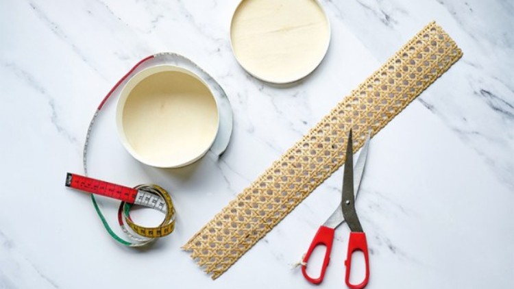 Eine rote Schere, ein Maßband, ein rundes Gefäß und ein gewebtes Band liegen auf einer weißen Marmoroberfläche.