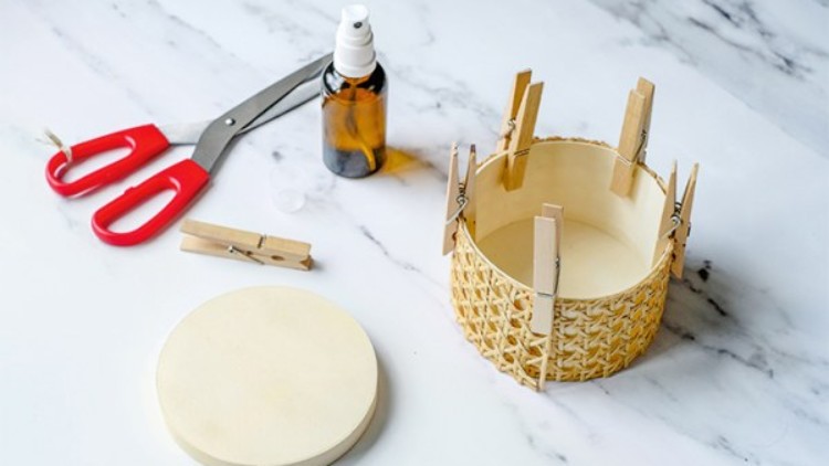 Runde Holzscheibe, Sprühflasche, Schere und Körbchen mit Wabenmuster, das mit Wäscheklammern fixiert ist, auf Marmortisch.