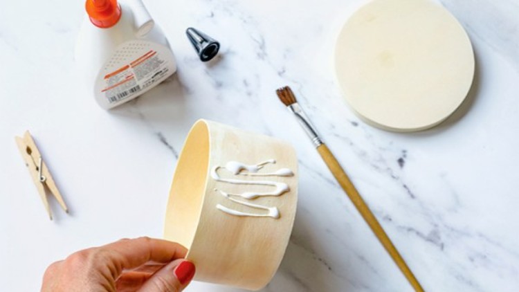 Eine Hand hält eine Holzdose mit Kleber darauf, daneben liegen Pinsel, Holzdeckel, Wäscheklammer, Tülle und Klebertube.