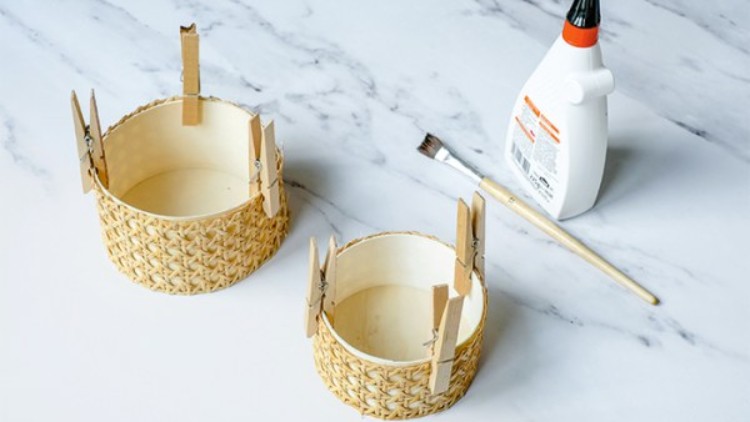 Zwei geflochtene Körbe werden mit Wäscheklammern fixiert, daneben stehen ein Pinsel und eine Flasche Bastelkleber.