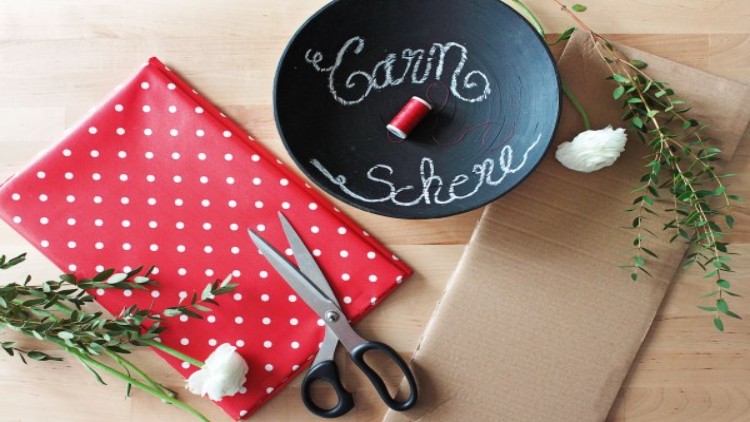 Rote Schere, rotes Garn, Stoff mit weißen Punkten, Zweige, weiße Blumen und Pappe auf einem Holztisch arrangiert.