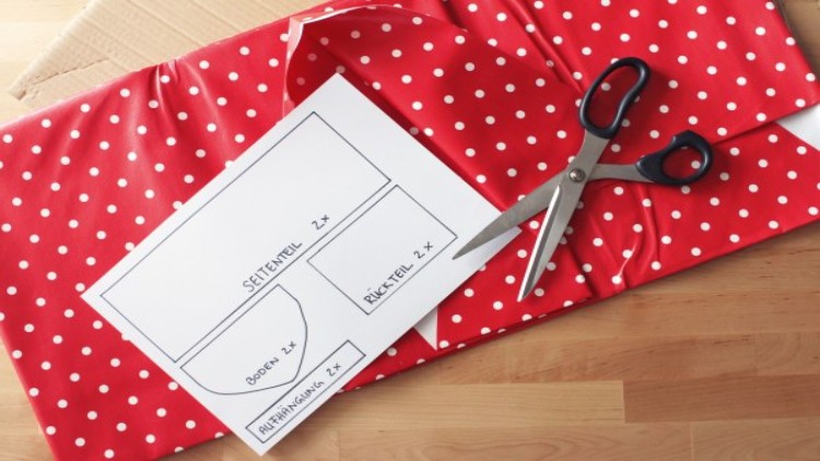 Ein weißes Papier mit Zuschneideanleitung liegt auf rotem Stoff mit weißen Punkten, daneben eine schwarze Schere.