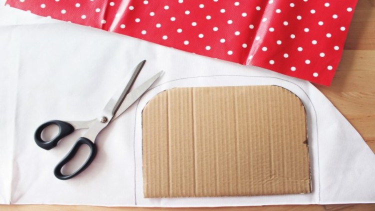 Eine Schere, ein rechteckiges Stück Pappe und rot-weiß gepunkteter Stoff liegen auf einer weißen Unterlage.