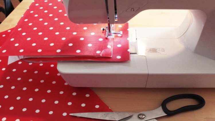 Eine weiße Nähmaschine näht roten Stoff mit weißen Punkten, daneben liegt eine schwarze Schere auf einem Holztisch.