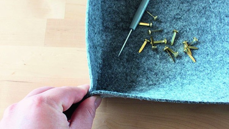 Eine Hand formt eine graue Filzschale, daneben liegen eine Ahle und goldfarbene Musterklammern auf hellem Holztisch.