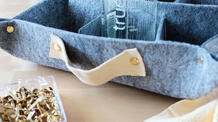 Grauer Filzkorb mit beigefarbenen Griffen und goldenen Nieten, daneben eine Plastikbox mit goldfarbenen Büroklammern.