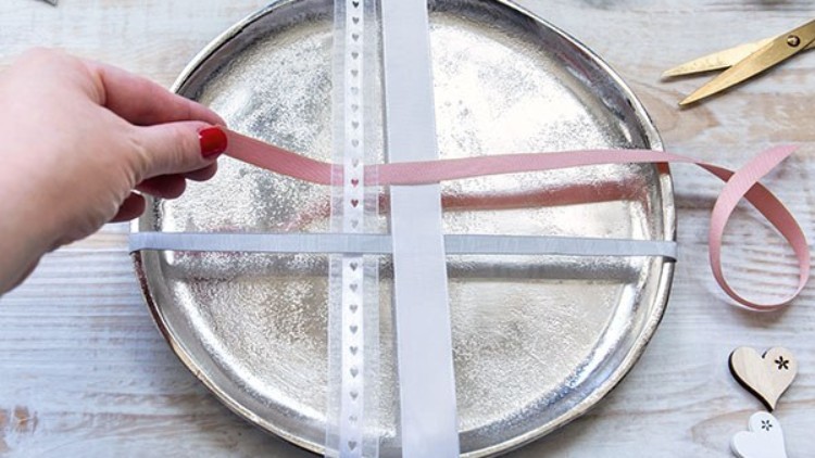 Eine silberne Platte wird mit weißen und rosa Stoffbändern verziert, eine Hand hält ein rosa Band fest.