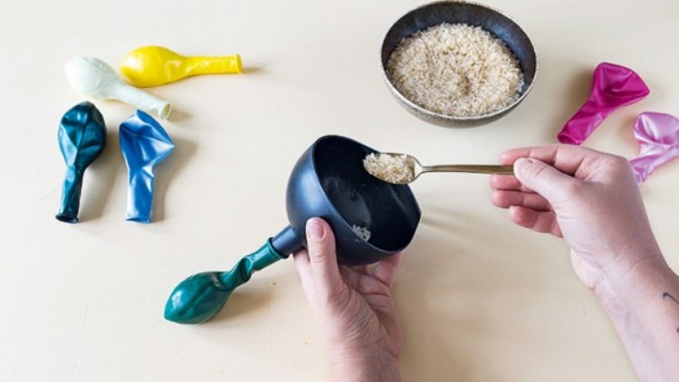 Zwei Hände befüllen mit einem Löffel einen schwarzen Trichter mit Reis, daneben liegen bunte Luftballons und eine Reisschale.