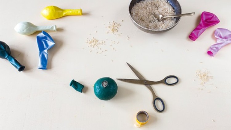 Auf einem Tisch liegen bunte Ballons, Schere, Klebeband, eine Schale Reis mit Löffel, Reisstreu und ein Ballon mit Reisfüllung.