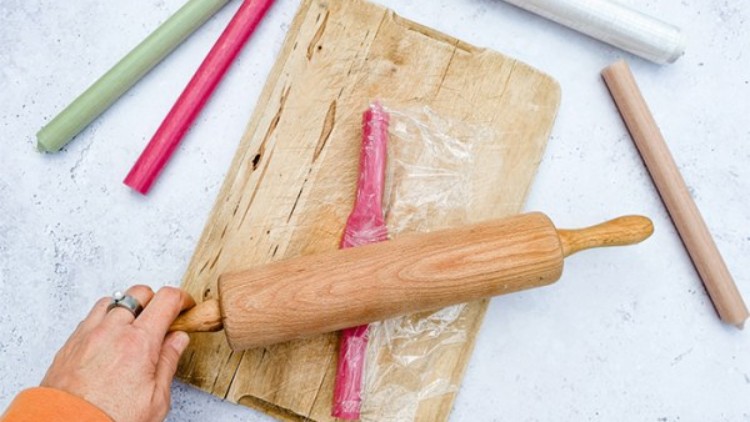 Eine Hand rollt mit einem Nudelholz eine pinke Masse in Frischhaltefolie auf einem Holzbrett aus, daneben liegen bunte Stangen.