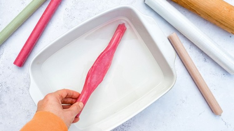 Eine Hand legt ein rotes Stück Rhabarber in eine weiße, rechteckige Auflaufform, umgeben von farbigen Nudelholzrollen.