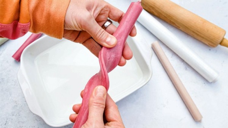 Hände formen eine gedrehte rosa Teigrolle über einer weißen Auflaufform, daneben liegen Nudelholz und andere Teigrollen.