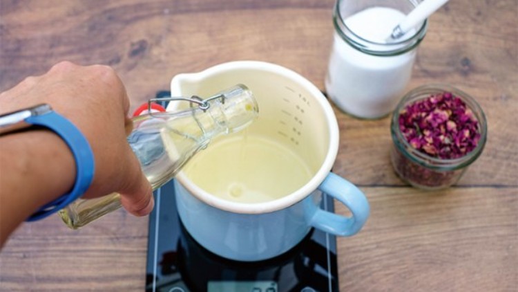 Eine Hand gießt Flüssigkeit aus einer Glasflasche in einen hellblauen Messbecher, daneben Salz und getrocknete Blüten in Gläsern.