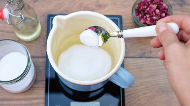 Ein weißer Löffel hält Zucker über einem blauen Topf auf einer Küchenwaage, umgeben von Glasgefäßen und getrockneten Blüten.