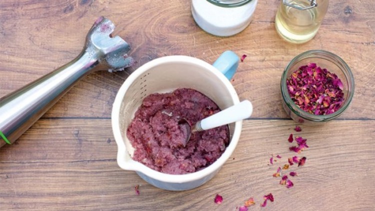 Ein Mixbehälter mit rosafarbener Masse, Pürierstab, Löffel, Glas mit Zucker, Ölflasche und getrockneten Rosenblättern auf Holztisch.